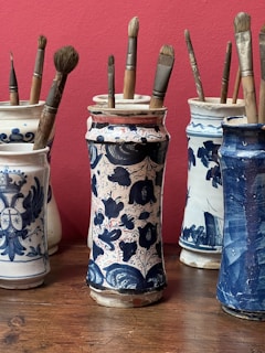Several ornate ceramic jars, each decorated with intricate blue patterns on a white background, hold collections of paintbrushes. The jars are arranged on a wooden surface against a red wall.