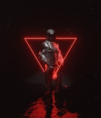 A person dressed in a reflective, futuristic outfit stands in front of a glowing red neon triangle. The reflective surface of the clothing contrasts with the dark background, and light reflects on a watery surface beneath.