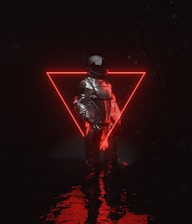 A person dressed in a reflective, futuristic outfit stands in front of a glowing red neon triangle. The reflective surface of the clothing contrasts with the dark background, and light reflects on a watery surface beneath.