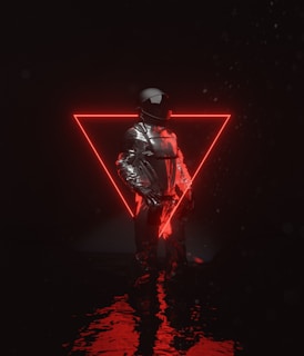 A person dressed in a reflective, futuristic outfit stands in front of a glowing red neon triangle. The reflective surface of the clothing contrasts with the dark background, and light reflects on a watery surface beneath.