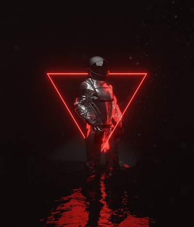 A person dressed in a reflective, futuristic outfit stands in front of a glowing red neon triangle. The reflective surface of the clothing contrasts with the dark background, and light reflects on a watery surface beneath.