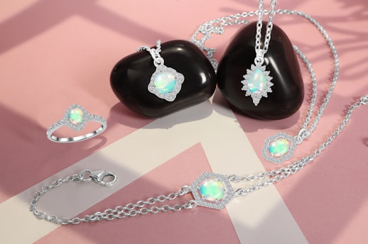 A collection of opal jewelry pieces, including rings, necklaces, and a bracelet, is elegantly arranged on a soft pink and beige background. The opals are luminous with iridescent colors, set in silver chains and surrounded by small crystals. Two smooth, black stones serve as a backdrop for some of the jewelry pieces, enhancing their shine.