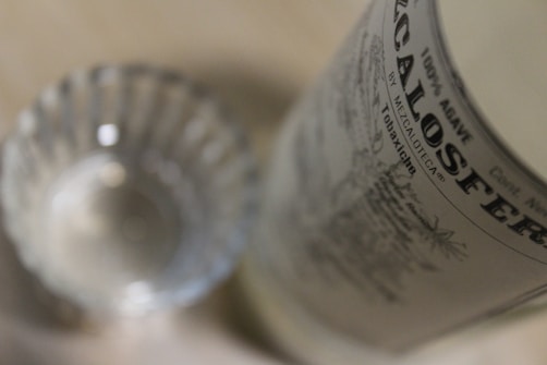 A close-up of a glass with intricate patterns alongside a partial view of a bottle with a label displaying text details. The focus is on the bottle, highlighting its branding.