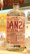 A clear glass bottle with a detailed label featuring the text 'Danza de los Pajaros'. The label includes various information such as alcohol content and origin. The bottle is surrounded by glass cups on a bar counter with a colorful, patterned background.