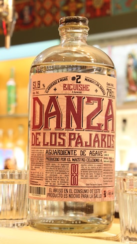 A clear glass bottle with a detailed label featuring the text 'Danza de los Pajaros'. The label includes various information such as alcohol content and origin. The bottle is surrounded by glass cups on a bar counter with a colorful, patterned background.