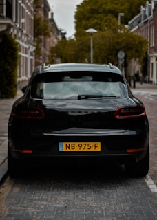 A black car is parked on a cobblestone street in a residential area. The buildings are traditional brick with white trim, and the trees are lush and green. The license plate on the car is yellow with black lettering.