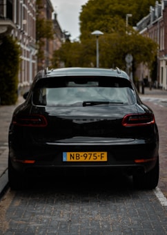 A black car is parked on a cobblestone street in a residential area. The buildings are traditional brick with white trim, and the trees are lush and green. The license plate on the car is yellow with black lettering.