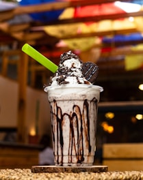 A tall, creamy milkshake topped with whipped cream, chocolate drizzle, and an Oreo cookie. A bright green straw is inserted into the shake. The background features blurred colorful umbrellas.
