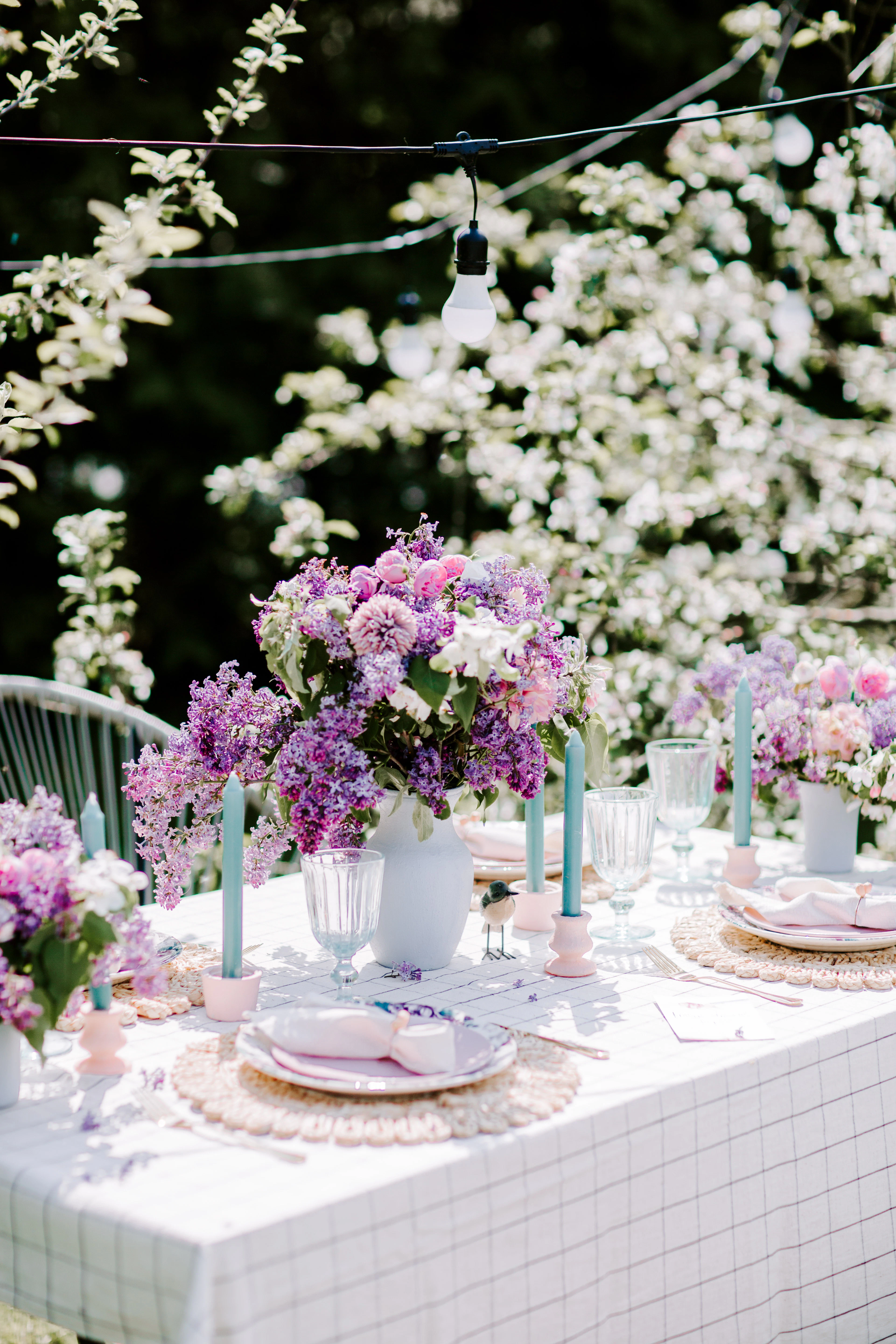 An outdoor table with lilac flower arrangements and pale blue taper candles beneath blossoming branches.