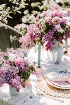 The image depicts a beautifully arranged outdoor table setting adorned with vibrant floral arrangements. There are two large vases filled with colorful flowers, predominantly in shades of purple and pink, accompanied by some green foliage. The table is set with elegant plates, cutlery, a napkin, and glassware. Blue taper candles are placed in pink candlesticks, adding to the aesthetic appeal. The background suggests a lush garden, providing a serene and refreshing atmosphere.