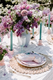 A beautifully arranged outdoor table setting features a centerpiece of vibrant purple and pink flowers in a vase, surrounded by teal candles. The table is covered with a white cloth and set with a woven placemat, floral-patterned plates, and a neatly folded cloth napkin with a ring. The scene is bright and elegant, suggesting a spring or summer gathering.