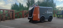 A robotic outdoor sweeper is positioned on a wooden deck surrounded by lush green trees. The machine is labeled with #ROBOCLEANING and appears to be designed for outdoor maintenance.