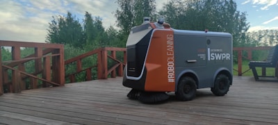 A robotic outdoor sweeper is positioned on a wooden deck surrounded by lush green trees. The machine is labeled with #ROBOCLEANING and appears to be designed for outdoor maintenance.