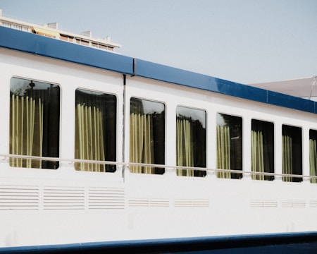 A large white vehicle, possibly a bus or train car, with a row of large windows framed by a blue trim. Light-colored curtains hang inside the windows, slightly drawn to allow sunlight to filter through. The overall appearance is clean and modern.