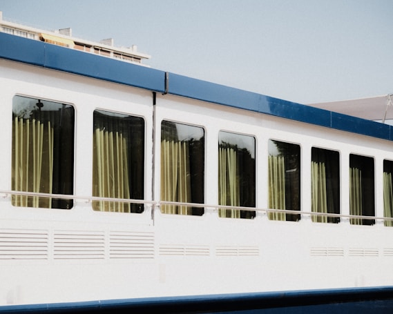 A large white vehicle, possibly a bus or train car, with a row of large windows framed by a blue trim. Light-colored curtains hang inside the windows, slightly drawn to allow sunlight to filter through. The overall appearance is clean and modern.