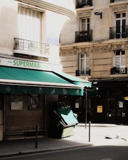 A street corner with a small supermarket that has a green awning extending over the sidewalk. There are a few posters on the wall next to the entrance. The store's name, 'Supermarché', is partially visible. The building has a slightly aged appearance with balconies and shuttered windows above the store. The scene appears to be set during daytime.