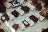 Various rocks and minerals are displayed in a museum showcase, each labeled with detailed information. The arrangement is neat, with small white cards providing the names and additional details of the specimens. The lighting is soft, giving the display an elegant and organized appearance.