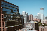 A vibrant cityscape featuring modern high-rise buildings with reflective glass windows. A mural adorns one of the lower structures, adding a touch of artistic flair. The clear blue sky contrasts with the urban architecture, creating a harmonious blend of nature and city life.