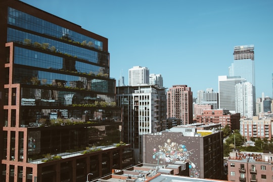 A vibrant cityscape featuring modern high-rise buildings with reflective glass windows. A mural adorns one of the lower structures, adding a touch of artistic flair. The clear blue sky contrasts with the urban architecture, creating a harmonious blend of nature and city life.