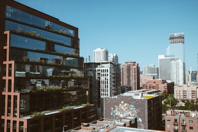 A vibrant cityscape featuring modern high-rise buildings with reflective glass windows. A mural adorns one of the lower structures, adding a touch of artistic flair. The clear blue sky contrasts with the urban architecture, creating a harmonious blend of nature and city life.