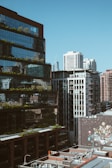 A cityscape featuring modern high-rise buildings with reflective glass facades and visible urban greenery. The foreground includes rooftops with multiple air conditioning units, while a large mural with various colorful designs is visible on a wall.