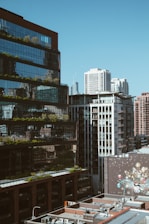 A cityscape featuring modern high-rise buildings with reflective glass facades and visible urban greenery. The foreground includes rooftops with multiple air conditioning units, while a large mural with various colorful designs is visible on a wall.