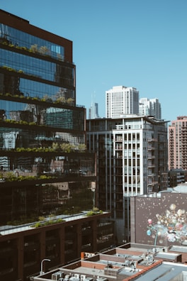 A cityscape featuring modern high-rise buildings with reflective glass facades and visible urban greenery. The foreground includes rooftops with multiple air conditioning units, while a large mural with various colorful designs is visible on a wall.