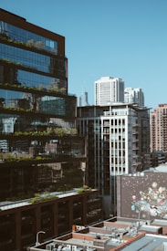 A cityscape featuring modern high-rise buildings with reflective glass facades and visible urban greenery. The foreground includes rooftops with multiple air conditioning units, while a large mural with various colorful designs is visible on a wall.