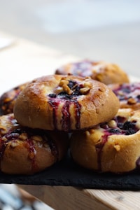 A close-up view of several freshly baked pastries placed on a dark surface. The pastries have a golden-brown crust with visible berry filling and a glossy finish. Crushed nuts are sprinkled on top, adding texture to the soft-looking pastries.