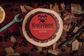 A red circular tin of beard balm from 'Daily Grind Beard Co.' is centered on a wooden slice with the text: 'Make moves or make excuses' and 'ALTUS'. Surrounding the tin are assorted natural elements including coffee beans, wood chips, a sliced orange, and a metal wrench on a rustic red surface.