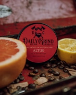 A round container of beard balm with a red label is surrounded by a cut grapefruit, a lemon, coffee beans, and wooden pieces, set against a rustic, textured background.