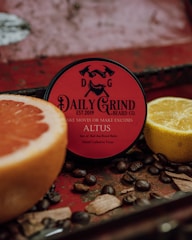 A round container of beard balm with a red label is surrounded by a cut grapefruit, a lemon, coffee beans, and wooden pieces, set against a rustic, textured background.