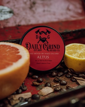 A round container of beard balm with a red label is surrounded by a cut grapefruit, a lemon, coffee beans, and wooden pieces, set against a rustic, textured background.