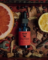 A bottle labeled 'Daily Grind ALTUS Quality Beard Oil' with a red and black design is surrounded by coffee beans, wood chips, a lemon slice, a grapefruit slice, and a small wrench on a red textured surface.