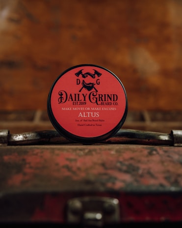 A round tin of beard balm with a red label is placed on a vintage, weathered suitcase. The branding on the label features the text 'DAILY GRIND BEARD CO.' with the slogan 'MAKE MOVES OR MAKE EXCUSES.' The background is blurred and has a warm, rustic tone.