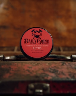 A round tin of beard balm with a red label is placed on a vintage, weathered suitcase. The branding on the label features the text 'DAILY GRIND BEARD CO.' with the slogan 'MAKE MOVES OR MAKE EXCUSES.' The background is blurred and has a warm, rustic tone.