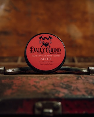 A round tin of beard balm with a red label is placed on a vintage, weathered suitcase. The branding on the label features the text 'DAILY GRIND BEARD CO.' with the slogan 'MAKE MOVES OR MAKE EXCUSES.' The background is blurred and has a warm, rustic tone.
