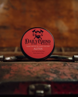 A round tin of beard balm with a red label is placed on a vintage, weathered suitcase. The branding on the label features the text 'DAILY GRIND BEARD CO.' with the slogan 'MAKE MOVES OR MAKE EXCUSES.' The background is blurred and has a warm, rustic tone.