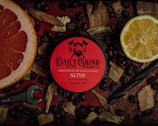 A tin of 'Daily Grind Beard Co.' beard balm is placed on a dark red surface surrounded by coffee beans, a half grapefruit, a half lemon, wooden shavings, and a wrench. The tin has a bold red label with black text and graphics, creating a masculine and rugged aesthetic.