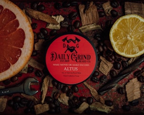 A tin of 'Daily Grind Beard Co.' beard balm is placed on a dark red surface surrounded by coffee beans, a half grapefruit, a half lemon, wooden shavings, and a wrench. The tin has a bold red label with black text and graphics, creating a masculine and rugged aesthetic.
