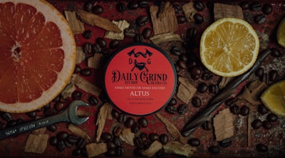 A circular container with red labeling from Daily Grind Beard Co. is surrounded by a variety of natural elements. There are coffee beans scattered around, as well as a grapefruit half and a lemon half placed on either side. Wooden chips add to the rustic aesthetic, and a wrench is visible at the bottom left.