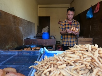 A person wearing a plaid shirt is handling several round pastries in a dimly lit room. The foreground features a large pile of fried snacks, possibly churros or a similar fried dough. Behind the person, cooking equipment and various plastic bags hang on the walls.