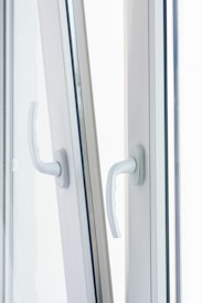 Two white window handles are positioned side by side on a slightly open window, catching soft, diffused daylight. The handles are modern in design, made of what appears to be plastic or painted metal.