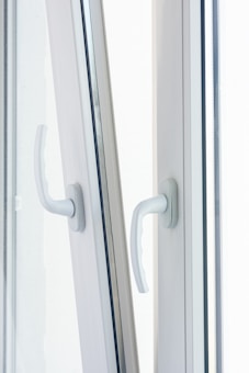 Two white window handles are positioned side by side on a slightly open window, catching soft, diffused daylight. The handles are modern in design, made of what appears to be plastic or painted metal.