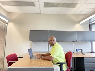A person is sitting at a wooden desk in an office setting, working on a laptop. The office has grey cabinets and a telephone on the desk. There is a red chair opposite the person, and large windows are letting in natural light.
