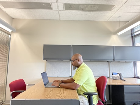 A person is sitting at a wooden desk in an office setting, working on a laptop. The office has grey cabinets and a telephone on the desk. There is a red chair opposite the person, and large windows are letting in natural light.