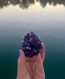 A hand with a lavender-colored nail polish holds a cluster of dark purple crystals against a tranquil lake background. The water reflects the soft colors of a setting or rising sun.