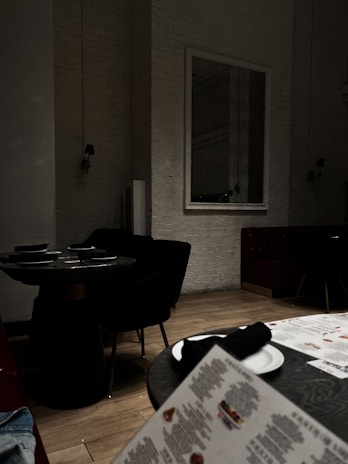 A dimly lit interior of a restaurant with dark furniture, including black chairs and round tables set with plates and folded napkins. A menu is visible in the foreground on a wooden floor. The walls are painted white with a large mirror reflecting part of the room.