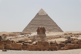 A large pyramid is set against a clear sky, with the famous Sphinx in the foreground. The scene includes several tourists walking on pathways around the historic site amid ancient stone ruins and structures, reflecting the grandeur of ancient Egyptian architecture.