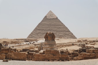 A large pyramid is set against a clear sky, with the famous Sphinx in the foreground. The scene includes several tourists walking on pathways around the historic site amid ancient stone ruins and structures, reflecting the grandeur of ancient Egyptian architecture.